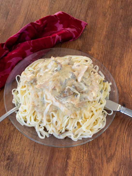 Easy chicken stroganoff with noodles in a clear bowl with a fork and knife near a red cloth napkin on a dark wood table