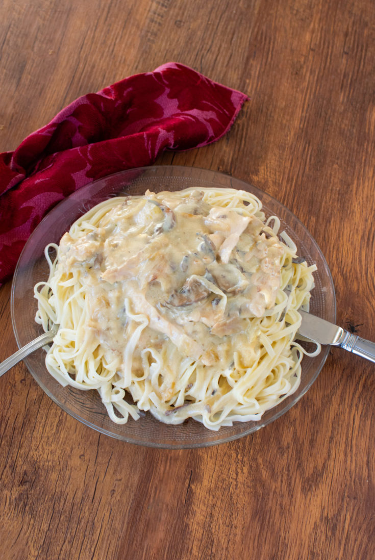 Easy chicken stroganoff with noodles in a clear bowl with a fork and knife near a red cloth napkin on a dark wood table