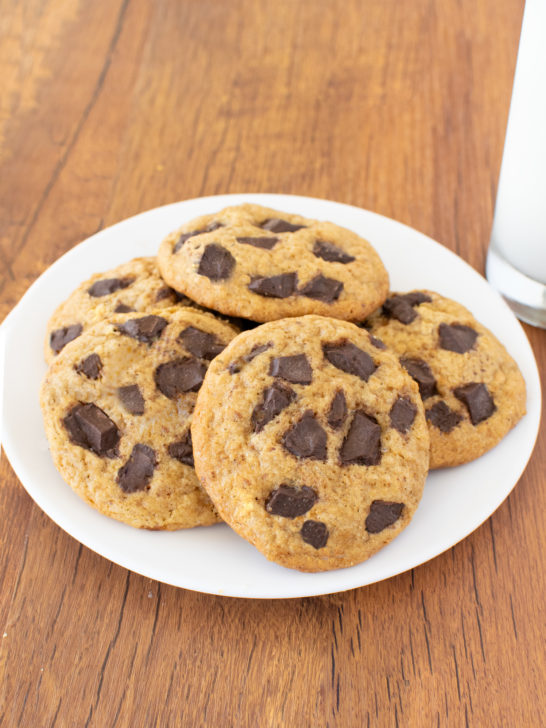 Easy Cakey Chocolate Chunk Cookies on a white plate with a clear glass of milk nearby on a dark wood table.