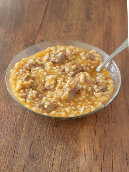 Easy Meat and Barley Soup in a clear glass bowl with a spoon on a dark wood table.