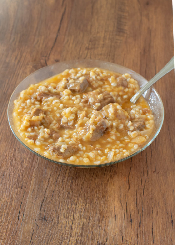 Easy Meat and Barley Soup in a clear glass bowl with a spoon on a dark wood table.