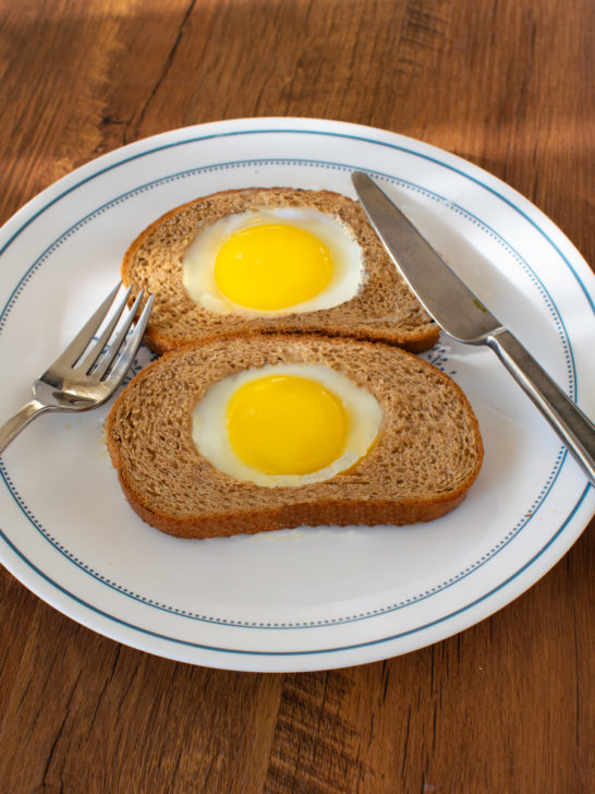 Two Easy eggs in a whole wheat hole on a white plate with a fork and knife on a dark wood table.