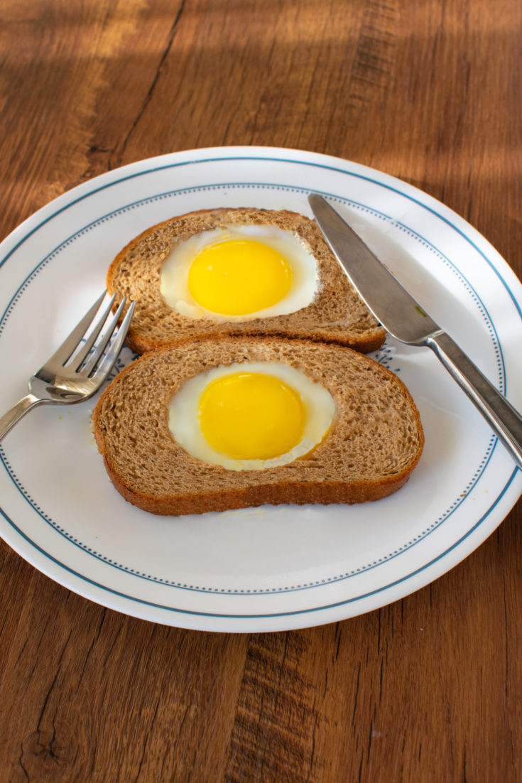 Two Easy eggs in a whole wheat hole on a white plate with a fork and knife on a dark wood table.