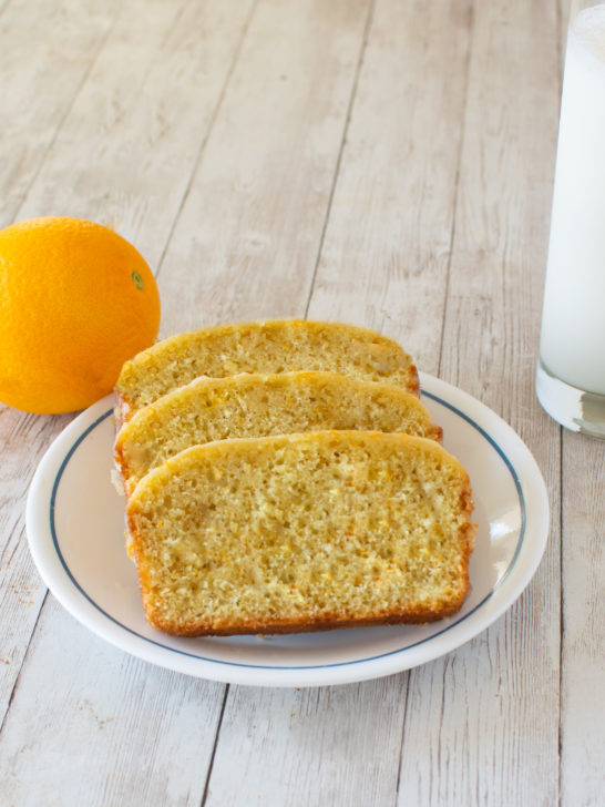 Easy Dairy-Free Orange Loaf Cake slices on a white plate with an orange and a clear glass of milk nearby on a white wood table